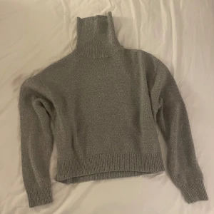 Grå stickad polotröja - Mysig grå stickad polotröja med hög krage och långa ärmar. Perfekt för kyliga dagar och ger en stilren look. Ribbstickade muddar vid ärmslut och nederkant för extra komfort.