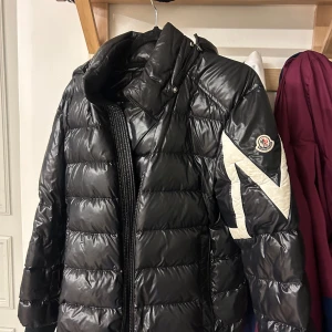 Svart dunjacka från Moncler - Snygg svart dunjacka från Moncler med ett stort vitt märke på ärmen. Jackan har en hög krage och dragkedja framtill. Perfekt för kyliga dagar. Ett litet hål på armen. Vid frågor så är det bara att skriva. Inga bud🙌