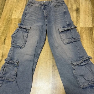 Grå cargobyxor med fickor - Säljer ett par grå cargobyxor med flera praktiska fickor på sidorna. Byxorna har en loose passform och är perfekta för en avslappnad stil. De är tillverkade i ett slitstarkt material och har en klassisk design med knapp och dragkedja. Priset går att diskuteras