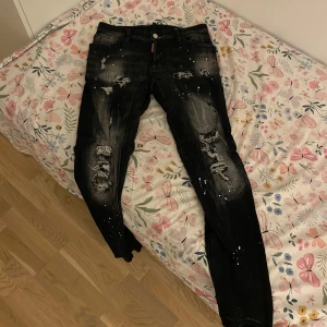 Svarta jeans med slitningar - Säljer ett par svarta jeans med coola slitningar och färgstänk. De har en knapp och dragkedja framtill samt fem fickor. Perfekta för en edgy look!