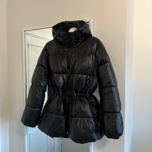 Svart pufferjacka  - Svart pufferjacka med huva och dragkedja. Jackan har justerbar passform i midjan. Jackan är i storlek M