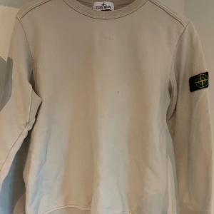 Stone Island Junior Crewneck  - Beige Stone Island Junior Crewneck i storlek 156. Det finns lite småfläckar på tröjan. (se bild) De är dock inte så synliga. Tveka inte på att skriva om ni har några frågor!