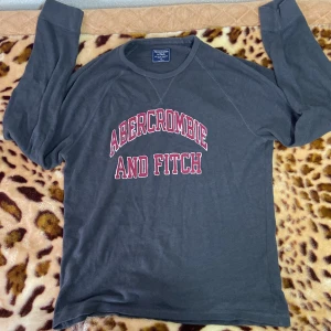 Grå långärmad tröja från Abercrombie & Fitch - Säljer en snygg grå långärmad tröja från Abercrombie & Fitch. Den har ett coolt tryck i rött på bröstet. Perfekt för höst och vinter, och den är superbekväm att ha på sig. Passar både till vardags och chillkvällar. 🖤