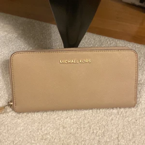 MICHAEL KORS plånbok - En stilren plånbok från Michael Kors. Köpt i Michael Kors butiken i Stockholm för många år sedan. Den är beige med tunna svarta detaljer runt om samt guld detaljer såsom kedjan runt om och inuti. Nypris 3430kr. Är fläckar på baksidan (sista bilden)