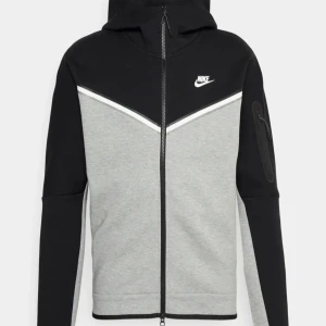 Nike tech fleece grå svart skriv för fler bilder - Säljer en snygg svart och grå hoodie från Nike med dragkedja framtill. Den har en cool design med vita detaljer och en praktisk ficka på ärmen. Perfekt för både träning och vardag. Stor Nike-logga på bröstet för extra stilpoäng!