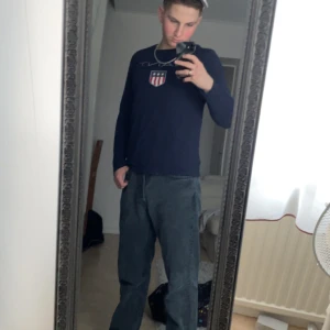 Mörkblå långärmad tröja med emblem - Säljer en snygg mörkblå långärmad tröja med ett coolt emblem på bröstet. Perfekt för höst och vår, och funkar både till vardags och lite mer uppklädda tillfällen. Tröjan har en normal passform och är superbekväm. Passar perfekt till jeans eller chinos!