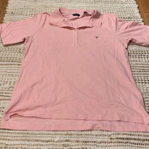 Rosa pikétröja från GANT - Säljer en snygg rosa pikétröja från GANT i storlek XL. Tröjan är i 100% bomull och har korta ärmar samt en klassisk krage med knappar. Perfekt för sommardagar eller en avslappnad stil. GANT-loggan är broderad på bröstet. Skicka ett meddelande vid intresse! 🌸