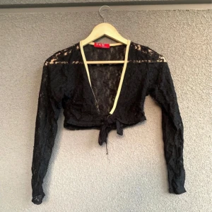 Svart spetsbolero från D&M - Säljer en elegant svart spetsbolero från D&M. Den är croppad och har långa ärmar med knytning framtill. Perfekt för en festlig kväll eller som ett stiligt komplement till din outfit. Använd den över en klänning eller topp för en chic look! ( Obs det gula tyget i fram har jag sprättat bort)