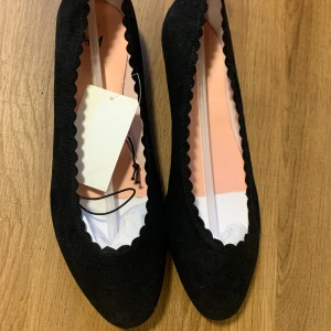 Svarta ballerinaskor från H&M - Säljer ett par eleganta svarta ballerinaskor från H&M i storlek 41. Skorna har en söt vågig kant och är i nytt skick. Perfekta för både vardag och festliga tillfällen. De är bekväma och stilrena, ett måste i garderoben! Storlek 41 men är mer som 40