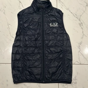 Svart dunväst från Emporio Armani - Säljer en snygg svart dunväst från Emporio Armani. Västen har en dragkedja framtill och är quiltad för extra värme. Perfekt för höst och vår när du vill hålla stilen utan att frysa. Den har en normal passform och EA7-loggan på bröstet. Passar både till vardags och fest!