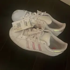 Säljer ett par vita Adidas Superstar i nytt skick. De har snygga glittriga detaljer i regnbågsfärger på sidorna och hälen. Perfekta för att ge din outfit lite extra bling! Skorna har klassisk snörning och är superbekväma. Passar perfekt för både vardag och fest. De har en pytte liten defekt som man ser på sista bilden men inget man märker av! Helt nya med lapp kvar! Jag säljer de pågrund av att de blev fel storlek och kvitto ej kan hittas. Kom gärna med bud! Köpta för 998 kr.