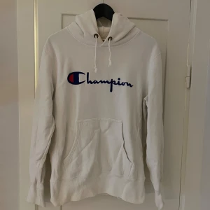 Vit hoodie från Champion - Säljer en snygg vit hoodie från Champion i storlek L. Den har en klassisk Champion-logga på bröstet och en liten logga på ärmen. Hoodien är i bra skick och perfekt för både höst och vinter. Den har en stor ficka fram och en bekväm huva med snören. Perfekt för en avslappnad stil!