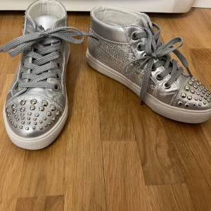 Silvriga sneakers med nitar från Oxygene - Säljer ett par coola silvriga sneakers från Oxygene. De har en glittrig yta med massor av nitar och paljett-detaljer på baksidan. Skorna har snörning och är i syntetmaterial. Perfekta för att sticka ut på festen!