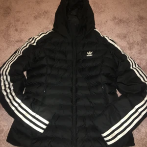 Svart dunjacka från Adidas stl 36 - Säljer en svart dunjacka från Adidas med de klassiska tre vita ränderna längs ärmarna. Jackan har en huva och dragkedja framtill. Perfekt för kyligare dagar och ger en sportig look. Adidas-loggan syns på bröstet. 🖤