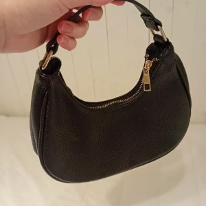 Svart handväska med dragkedja - Snygg svart handväska i skinn med guldfärgad dragkedja. Perfekt för att bära dina nödvändigheter med stil. Den har en kort rem som gör den enkel att bära i handen.