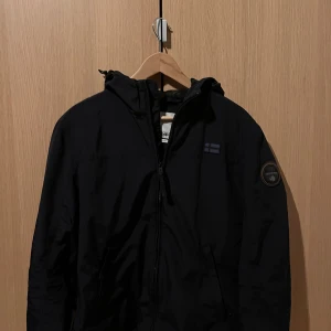 Svart jacka från Napapijri - Säljer en svart jacka från Napapijri med dragkedja och huva. Jackan har en broderad logotyp på ärmen och en liten flagga på bröstet. Perfekt för höst och vår. Kom med frågor!