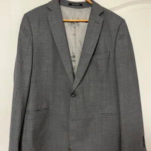 Grå kavaj från Oscar Jacobson - Snygg grå kavaj från Oscar Jacobson med klassisk passform. Kavajen har två knappar framtill och en stilren design. Perfekt för både formella och semi-formella tillfällen.