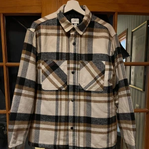 Rutig overshirt i beige och svart - Säljer en snygg ”helt ny” rutig overshirt i beige, svart och brunt. Lätt vadderad inuti. Den har långa ärmar och knappar framtill. Två stora bröstfickor med lock ger en cool look. Perfekt för lager-på-lager-stil. Från djur/rökfritt hem. 