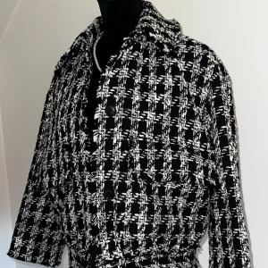Svartvit rutig kort kappa - Snygg svartvit rutig kappa med knappar och framfickor. Den ger en stilren och elegant look, perfekt för alla tillfällen. Med sin klassiska krage och långa ärmar är den idealisk för kyligare dagar. En fantastisk tillägg till din garderob!