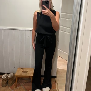 Svart byxdress med knytband - Snygg svart byxdress med hög midja och knytband i midjan. Byxdressen har en ärmlös design och vida ben som ger en elegant look. Perfekt för en stilren outfit.