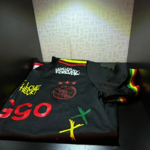 Svart Ajax fotbollströja med reggae-tema - Cool svar tajax  fotbollströja med reggae-inspirerade detaljer och färger. Texten 'Legends Forever' och 'Live the Life' pryder tröjan tillsammans med ett stiliserat ansikte. Ärmarna har ränder i rött, gult och grönt.