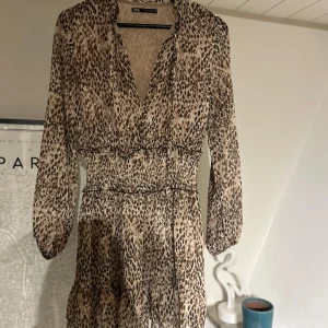 Leopardmönstrad klänning från Zara - Trendig leopardmönstrad klänning från Zara med v-ringning och långa ärmar. Klänningen har en smickrande passform med elastisk midja och volangdetaljer. Perfekt för att sticka ut med stil.