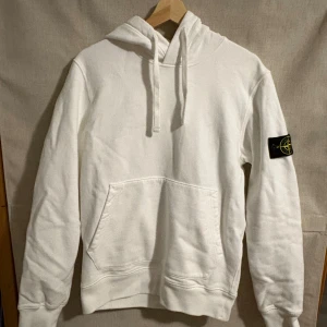 Vit hoodie från Stone Island - Säljer en stilren vit hoodie från Stone Island med klassisk patch på ärmen. Tröjan har en känguruficka och justerbar huva med snören. Perfekt för en avslappnad look.