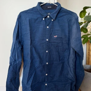 Mörkblå skjorta från Hollister - Säljer en stilren mörkblå skjorta från Hollister i slim fit. Skjortan har långa ärmar och knappar framtill. Den är tillverkad i ett stretchigt material för extra komfort. Perfekt för en avslappnad men ändå uppklädd look.