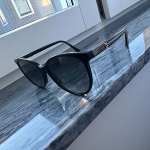 Svarta solglasögon från Gucci - Snygga svarta solglasögon från Gucci med en elegant cat-eye design. Bågarna har en diskret logga på sidan och är tillverkade i ett glansigt material. Perfekta för att ge en touch av lyx till din stil.