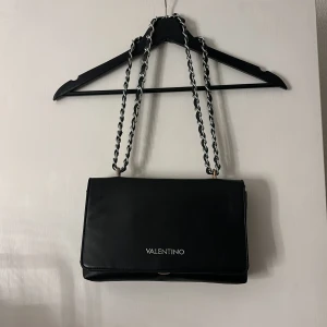 Svart axelväska från Valentino - Snygg svart axelväska från Valentino med kedjedetaljer på axelremmen. Väskan har en stilren design med logotyp framtill. Perfekt för att ge en elegant touch till din outfit.