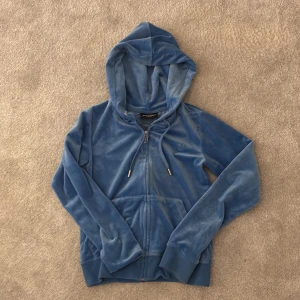 Blå velourjacka från Juicy Couture - Säljer en snygg blå velourjacka från Juicy Couture med huva och dragkedja. Jackan har ett broderat JC-märke på bröstet och ribbade muddar. Perfekt för en avslappnad stil.