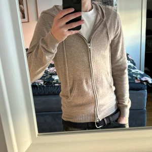 Beige cashmere hoodie från John Henric - Säljer en lyxig beige hoodie från John Henric i mjuk kashmir. Tröjan har dragkedja framtill och en bekväm huva med snörning. Perfekt för en stilren och avslappnad look.
