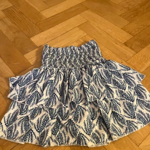 Vit och blå kjol med mönster - Charmig kortkjol i vitt med blått mönster. Kjolen har en elastisk midja för bekväm passform och är fodrad för extra komfort. Perfekt för varma dagar.