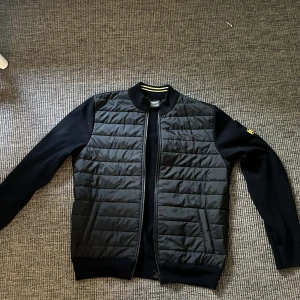 Svart quiltad jacka från Barbour - Snygg svart quiltad jacka från barbour  med dragkedja och ribbad krage. Jackan har långa ärmar och en stilren design med ett märke på ärmen. Perfekt för en casual look.