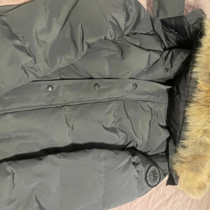 Grå dunjacka från Canada Goose - Säljer en grå dunjacka från Canada Goose med äkta pälsdetaljer på huvan. Jackan har en klassisk design med knappar och dragkedja framtill samt en broderad logotyp på ärmen. Perfekt för kalla vinterdagar.
