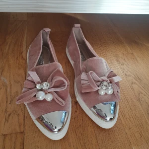 Rosa loafers med pärldetaljer - Snygga rosa loafers i sammet med dekorativa rosetter och pärlor framtill. Skorna har en metallisk spetsig tå och en vit sula. Perfekta för att ge en touch av glamour till din outfit. Använda 1 gång bara.