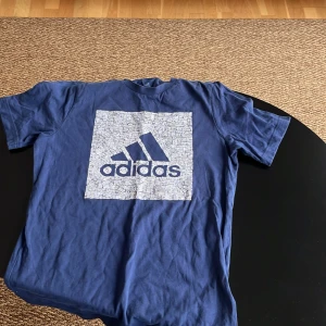 Blå t-shirt från Adidas - Säljer en blå t-shirt från Adidas med ett stort vitt och blått logotryck på framsidan. Perfekt för en sportig look eller avslappnad stil. T-shirten är kortärmad och gjord i mjukt bomullsmaterial.