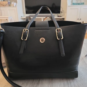 Svart axelväska från Tommy Hilfiger - Snygg svart axelväska från Tommy Hilfiger med guldfärgade detaljer och dubbla handtag. Väskan har en stilren design med ett rymligt inre och en praktisk axelrem. Perfekt för att bära med sig allt man behöver under dagen.