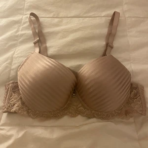 Rosa bh med spetsdetaljer - Säljer en rosa pushup bh med ränder och spetsdetalj längs kanten. Den har justerbara axelband och en liten hjärtformad dekoration framtill. 