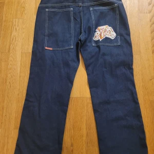 Mörkblå jeans från JNCO - Säljer ett par mörkblå jeans från JNCO med unika detaljer som en broderad tärning på bakfickan och en siffra 7 på framsidan. Jeansen har en loose passform och är perfekta för en avslappnad stil.
