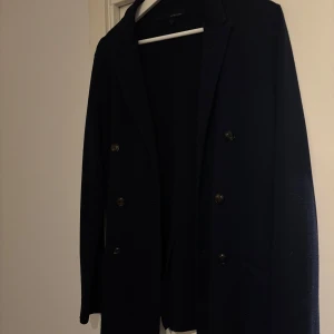 Mörkblå cardigan från Lardini - Säljer en elegant mörkblå cardigan från Lardini i 100% ull. Kavajen har en klassisk design med dubbelknäppning och långa ärmar. Perfekt för en stilren look.