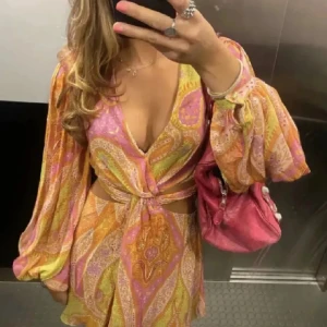 Färgglad kortklänning med knyt - Säljer en färgglad kortklänning med ett livfullt mönster i rosa, orange och gult. Klänningen har en djup v-ringning och långa ärmar med resår vid ärmsluten. Den har en knytning framtill som ger en snygg detalj och en luftig passform.