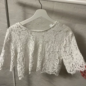 Vit spetsblus - Säljer en söt vit spetsblus med trekvartsärm. Blusen är croppad och har ett vackert blommigt mönster. Perfekt för en somrig look!
