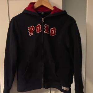 Svart hoodie från Polo Ralph Lauren - Säljer en svart hoodie från Polo Ralph Lauren med röd insida i huvan och texten 'POLO' i rött och vitt på bröstet. Tröjan har dragkedja framtill och långa ärmar. Perfekt för en avslappnad stil.