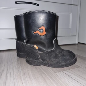 Svarta boots med orange detalj - Säljer ett par svarta vinterstövlar från kavat i stl 25 och är i skinn och med gummifot, med en cool orange logga på sidan. De har en robust design och är i använt skick. Perfekta för höst och vinter med sin slitstarka sula och bekväma passform. De har också en praktisk flik upptill för enkel påtagning.