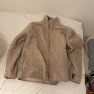 Beige teddyjacka med dragkedja - Mysig beige teddyjacka med dragkedja och hög krage. Perfekt för kyliga dagar med sin mjuka och varma känsla. Jackan har långa ärmar och praktiska tumhål för extra komfort.