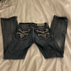 Rock Revival jeans - Snygga mörkblå jeans från Rock Revival med unika broderade detaljer på bakfickorna. Jeansen har en lågmidjad passform och är dekorerade med kontrasterande vita sömmar. Dom är 37cm i midjan och 78cm i inner benlängd. Jag säljer dom eftersom dom har blivit förstora. Dom har slitningar vid hälarna eftersom dom var för långa, men det finns inga andra slitningar. Kontakta för mer information.