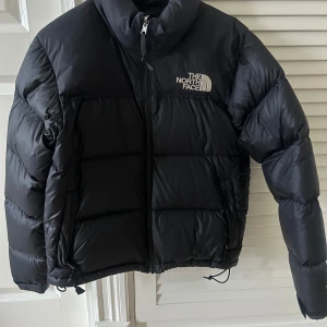 Svart dunjacka från The North Face - Säljer en svart dunjacka från The North Face. Jackan har en dragkedja framtill och en hög krage för extra värme. Den är perfekt för kalla dagar och har en stilren design med The North Face-logga på bröstet och ryggen.    700 nuptse