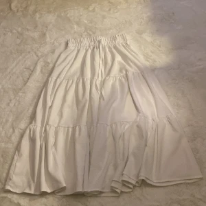 Vit långkjol från Shein - Säljer en vit långkjol från Shein med elastisk midja och knytband. Kjolen har en luftig design med flera volanger som ger en fin rörelse. Perfekt för varma dagar och en stilren look.