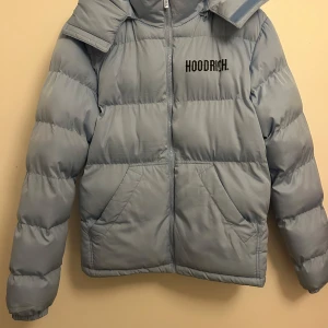 Ljusblå pufferjacka från Hoodrich - Säljer en snygg ljusblå pufferjacka från Hoodrich med stor logga på bröstet och ryggen. Jackan har en dragkedja framtill och är perfekt för kyligare dagar. Den har långa ärmar och en mysig huva.